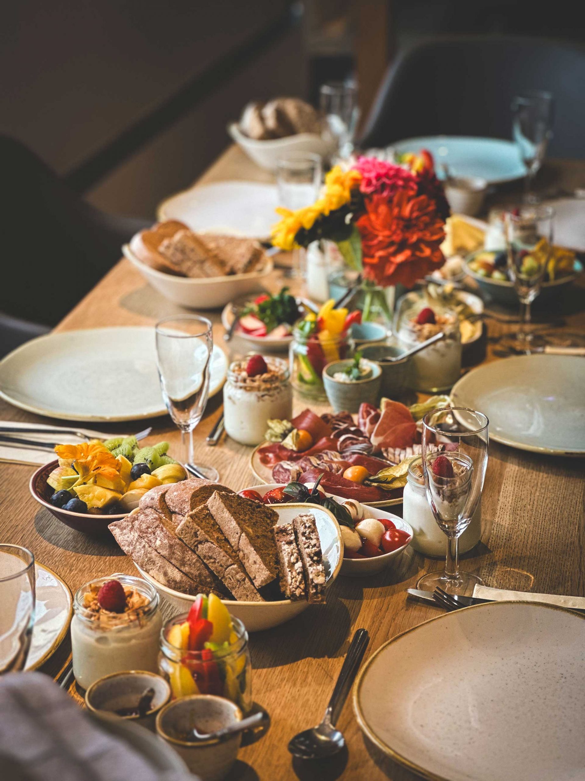 Hübsch gedeckter Frühstückstisch mit Blumen, Joghurt, Brot, Wurstplatte, Gemüse, Obst, Antipasti