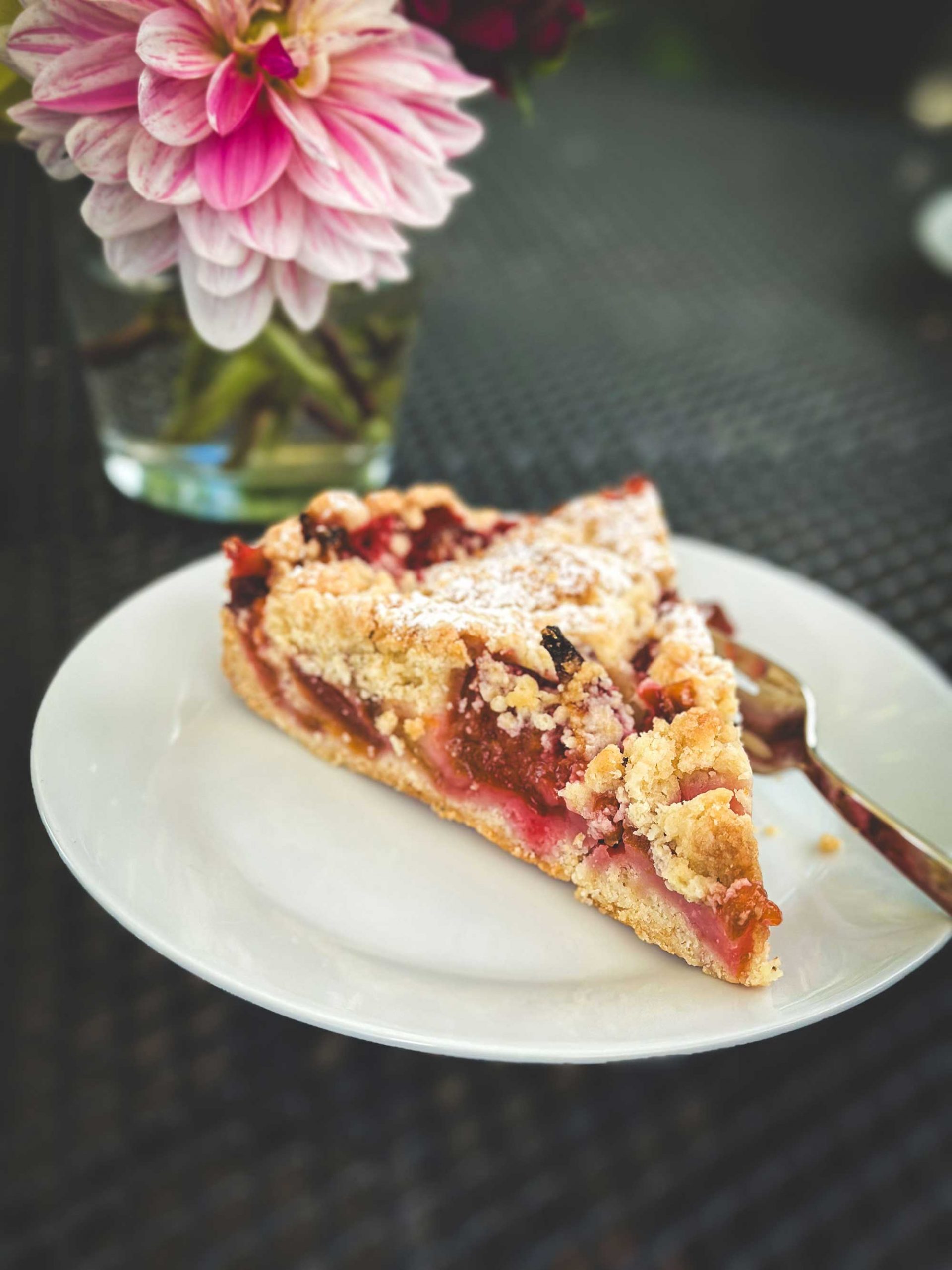 Ein Stück Zwetschgenkuchen mit Streusel auf einem Teller mit einer Gabel und einem Blumenstrauß daneben